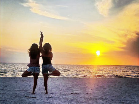 Two women practicing yoga on the beach at sunset, standing in tree pose, with the ocean and golden sky in the background.