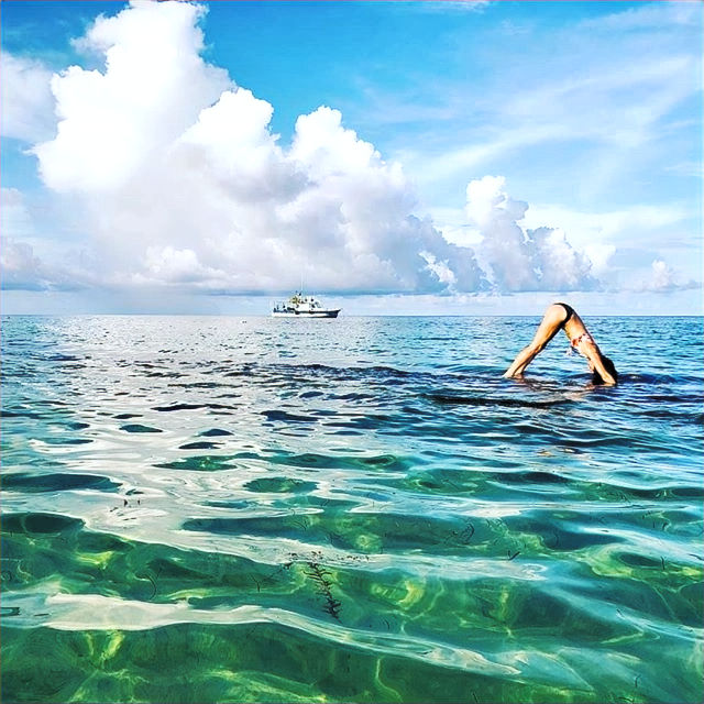 Person practicing yoga on clear blue ocean water, forming a bridge pose. A ship is visible on the horizon under a sky with fluffy clouds.