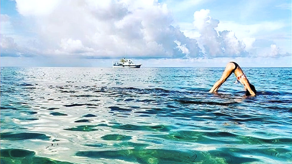 Person practicing yoga on clear blue ocean water, forming a bridge pose. A ship is visible on the horizon under a sky with fluffy clouds.