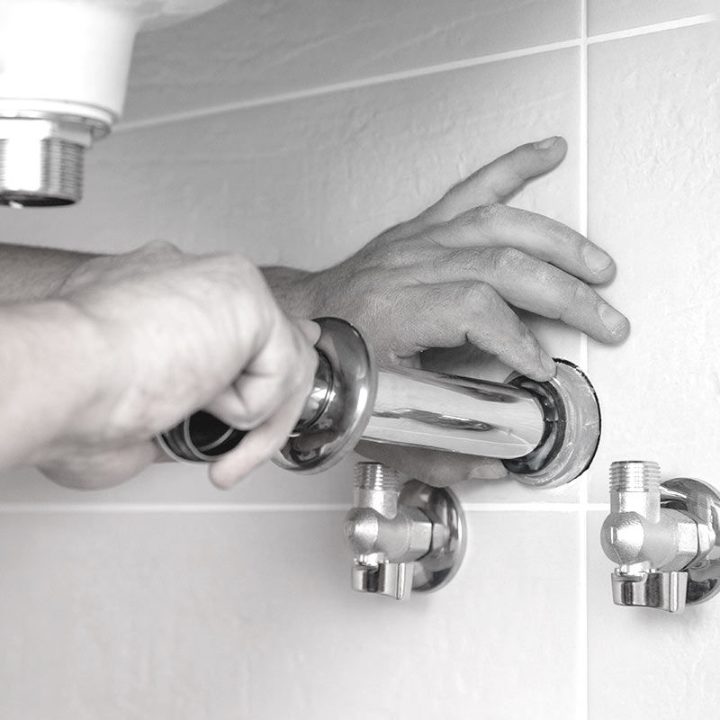 Two hands of plumber near wall with white tiles. One hand is leaning against the tiles, while other is installing chrome faucet.