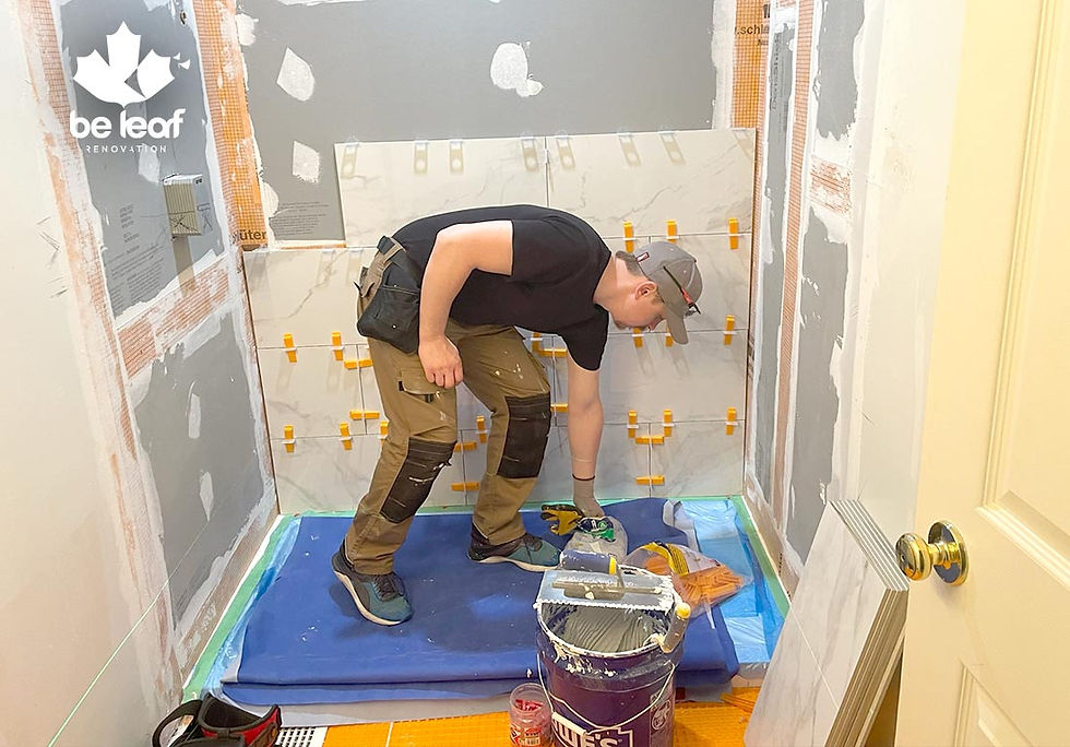 Man in a cap lays tiles in a small room using spacers. Blue tarp and bucket on the floor. Gray walls with white patches. Logo: be leaf renovation.