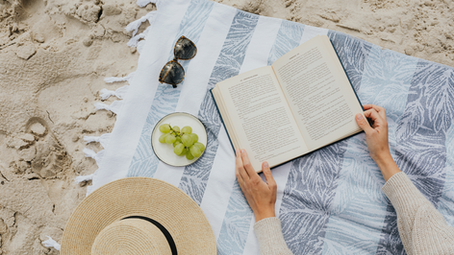 A woman sits on a blue beach blanket on the sand reading a book. On the blanket are a plate of green grapes, a sunhat, and sunglasses.