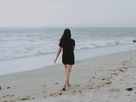 A woman in black shorts and a t-shirt and tennis shoes walks along a beach as the waves roll in.