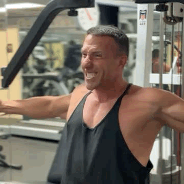 Theo Marcopoulos homme musclé en débardeur noir, bras écartés sur une machine de musculation, souriant dans une salle de sport.