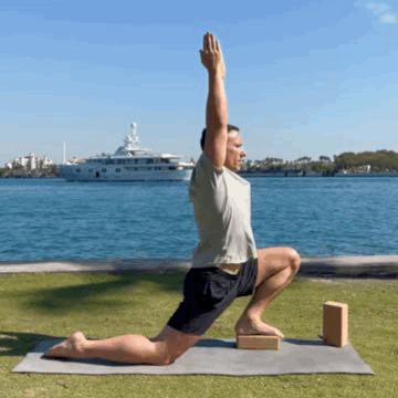 Theo Marcopoulos is practicing an outdoor yoga session by the water, performing a low lunge pose