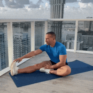 Man in a blue shirt stretches on a blue mat on a rooftop terrace with city buildings in the background.