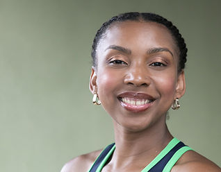 A close-up portrait of a woman with braided hair pulled back, smiling warmly at the camera. She wears small gold hoop earrings and a navy athletic dress with green trim. Her expression is open and confident, set against a soft green studio backdrop.