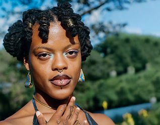 A close up portrait of a brown skin person outdoors with soft greenery and sky in the background. The person is wearing a blue top and their gaze meets the camera directly with a tattooed hand near their collarbone and neck. Their locs are twisted in the front with gold adornments and two buns in the back.