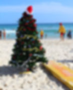 Christmas tree with Santa hat on a sunny beach; people swimming; lifeguard in yellow; vibrant ocean backdrop; festive and playful mood.
