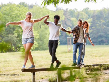 Friends help each other balance while walking on a wooden beam outdoors.
