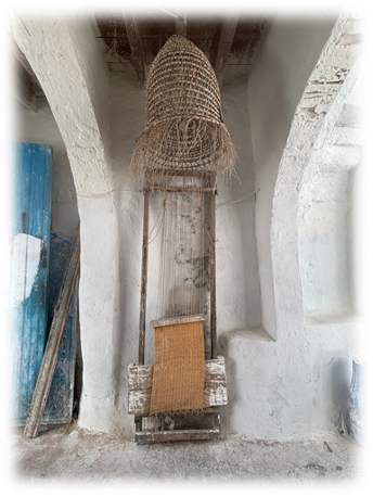 Houmt souk: Vieille ruche en osier suspendue à un mur blanc sous des arcs. Porte bleue patinée à gauche, ambiance rustique et paisible.