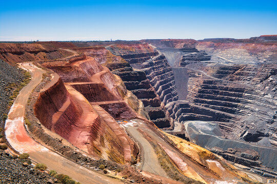 Open-pit mine with layered rock formations in red and gray. Dirt roads wind through the excavation. Clear blue sky in the background.