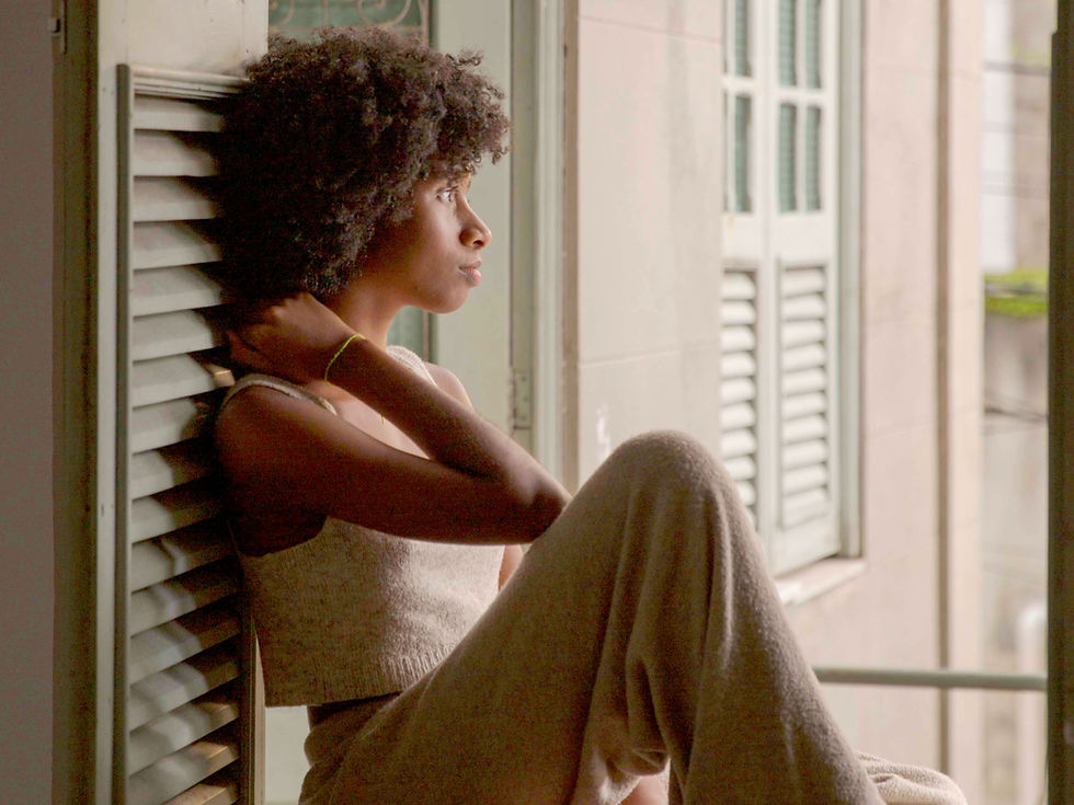 A young woman with natural curly hair sits indoors by a window, leaning against the wall and looking outside with a thoughtful, distant expression. Her posture and gaze suggest quiet reflection or sadness.