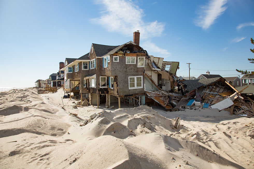A beachfront home severely damaged by a coastal storm, with sand piled high around the structure and parts of the building collapsed under the force of wind and waves.
