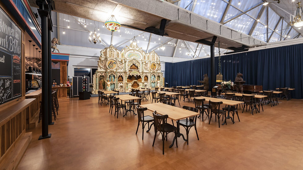 Ein leerer Saal mit Holztischen, Stühlen und einer großen Orgel im Hintergrund. Dunkelblaue Vorhänge und ein bunter Kronleuchter.