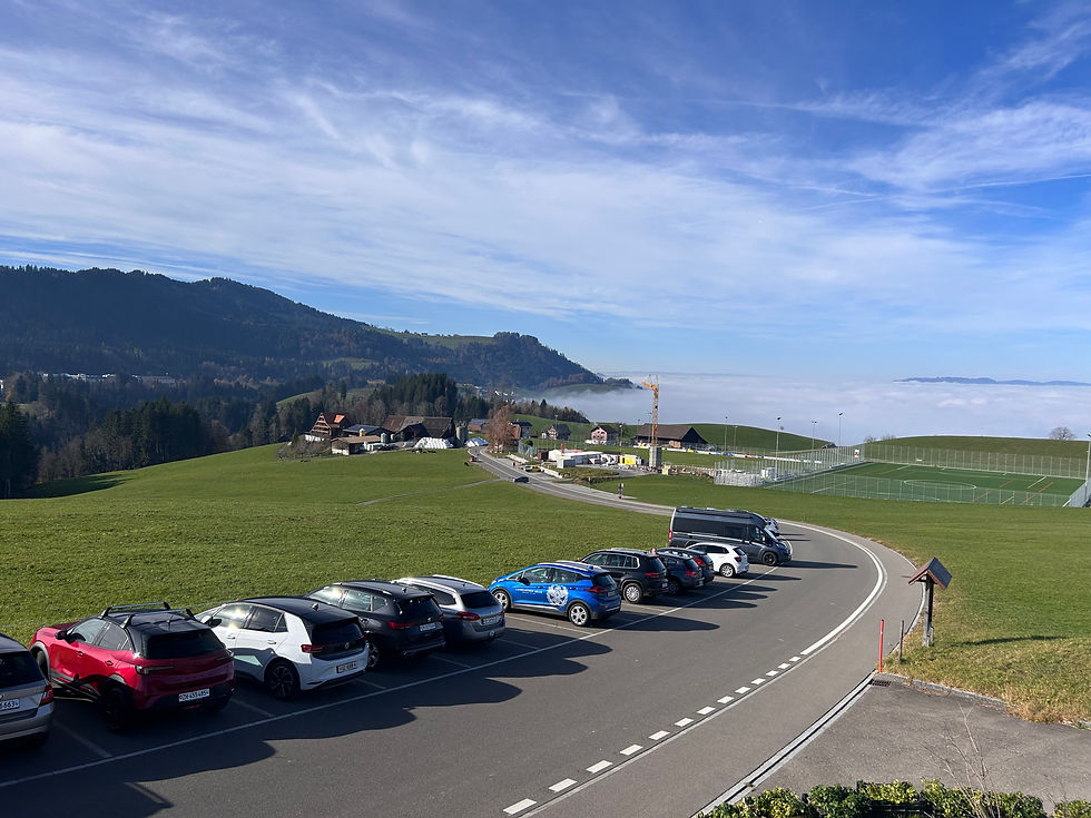 Autos parken neben einer Straße in einer grünen, hügeligen Landschaft mit blauem Himmel und Wolkendecke im Hintergrund.