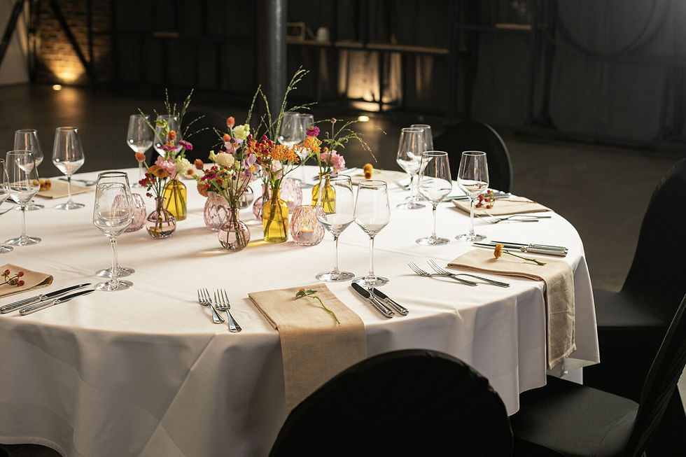 Elegant gedeckter runder Tisch mit weißen Tischdecken, bunten Blumen in Vasen, Weingläsern und Besteck, in einem dimm beleuchteten Raum.