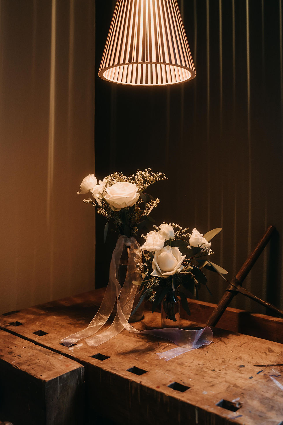 Weiße Rosen in Glasvasen auf Holztisch unter warmer, gestreifter Lampe. Gemütliche Atmosphäre mit Schatten und zarten Bändern.