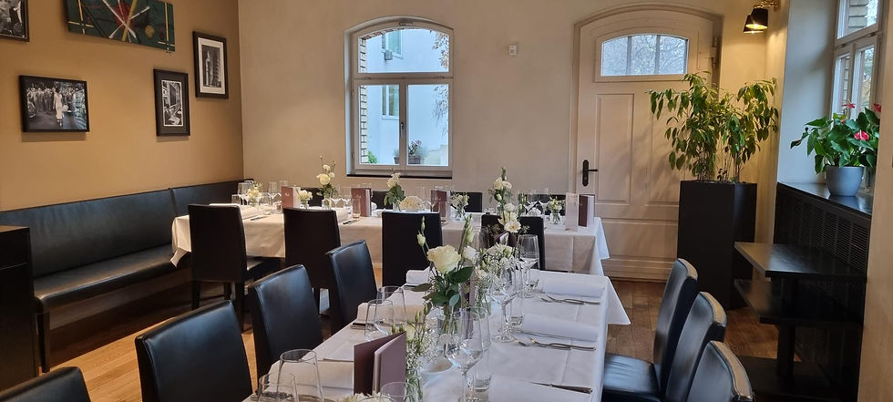 Elegantes Esszimmer mit gedeckten Tischen, weißen Tischdecken, Blumenarrangements und dekorativen Bildern an beige Wänden. Fenster im Hintergrund.