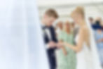 Brautpaar tauscht Ringe in heller Hochzeitsumgebung. Braut lächelt, Gäste im Hintergrund. Festliche Stimmung, weiße und grüne Kleidung.