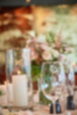 Kerzen und ein Blumenstrauß in Pastellfarben sind in Glasvasen auf einem dekorierten Tisch arrangiert. Im Hintergrund sanftes Bokeh.