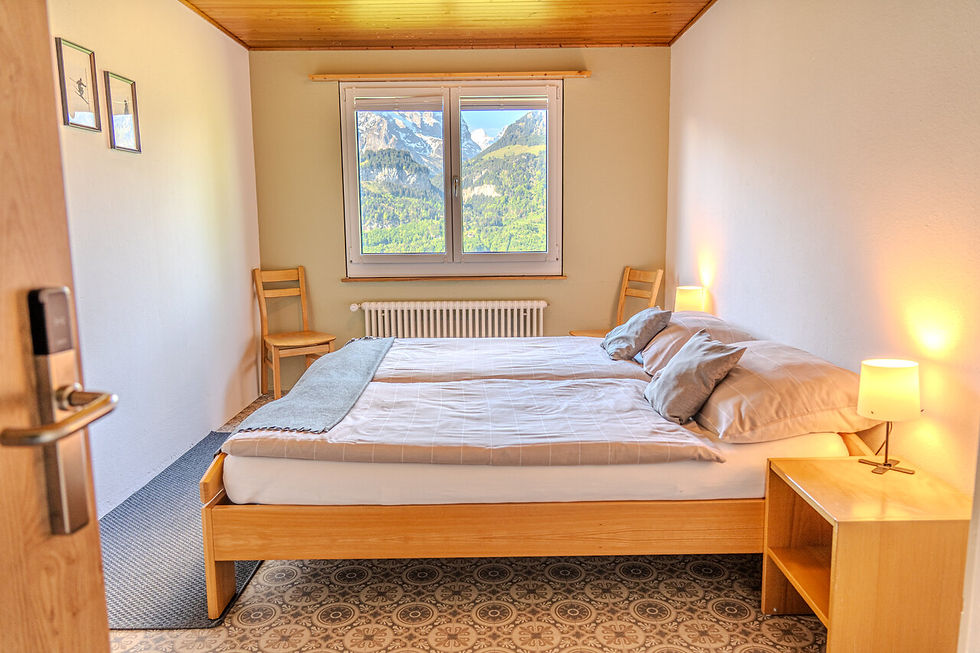Gemütliches Schlafzimmer mit Doppelbett, zwei Stühlen und Nachttischlampen. Fenster mit Bergblick, helle Wände, graue Decke.