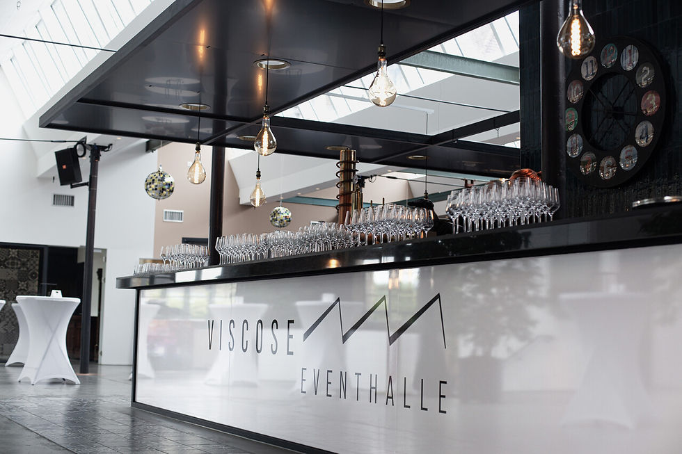 Moderne Bar in heller Eventhalle. Zahlreiche Weingläser auf Theke, Hängelampen, Uhr im Hintergrund. Text: "Viscose Eventhalle".