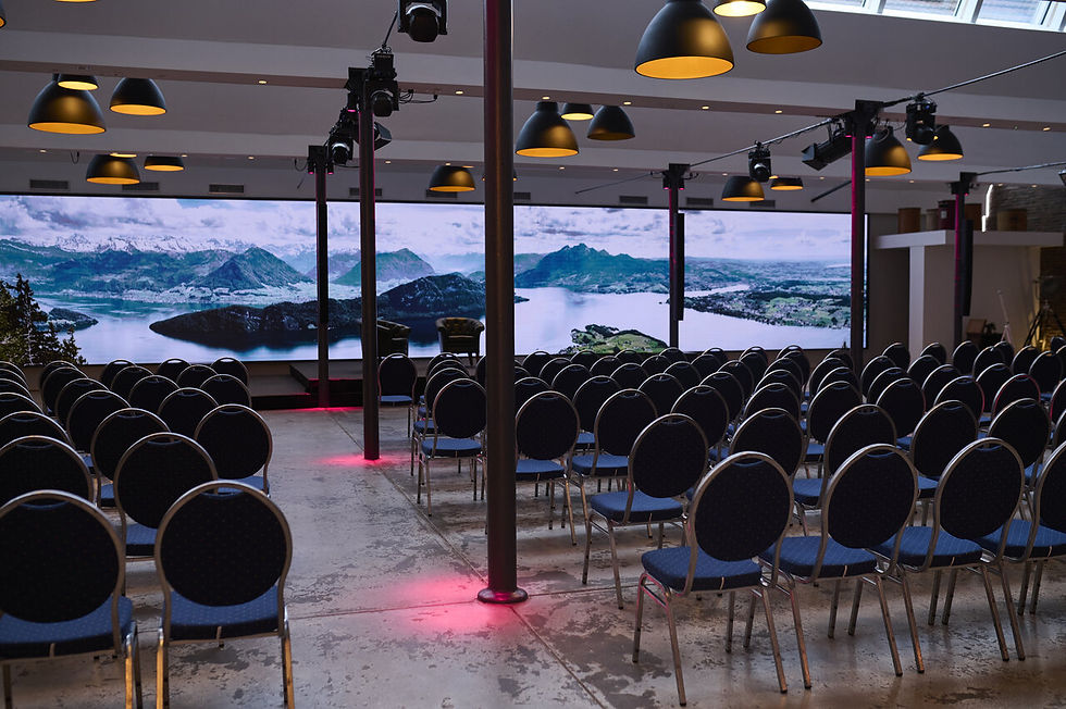 Stuhlreihen in modernem Konferenzraum, riesiger Bildschirm zeigt Seelandschaft. Dunkle Lampen hängen von der Decke, rote Lichter am Boden.
