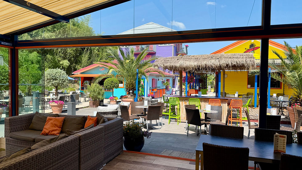 Bunte Strandbar mit Palmen, bunten Stühlen und Hütten. Gemütliche Sofas im Vordergrund. Sonniges Wetter, entspannte Atmosphäre.
