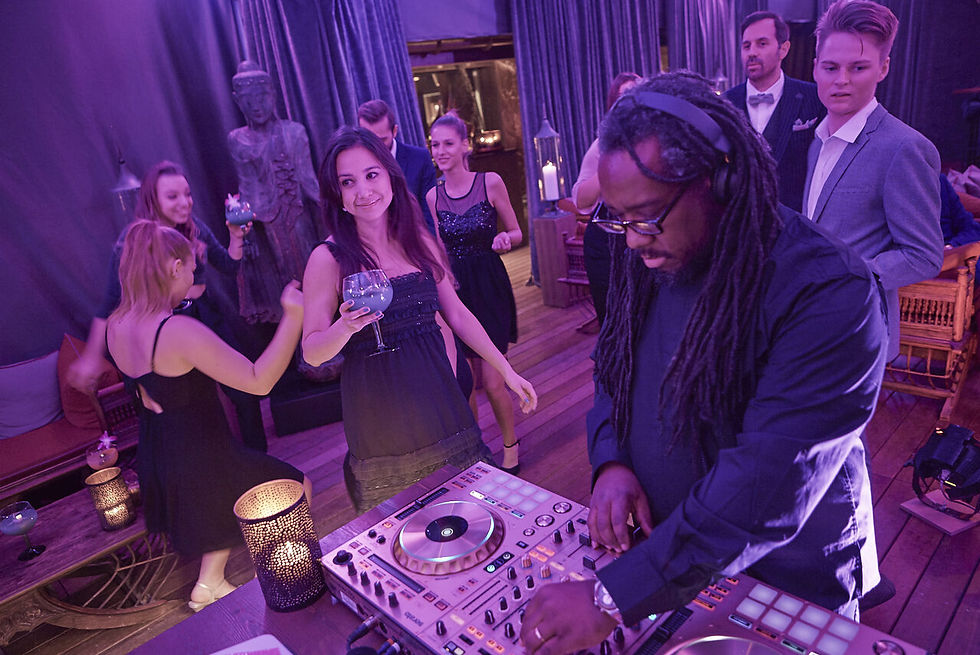 DJ legt auf Party auf. Menschen in eleganter Kleidung tanzen. Lila Licht, entspannte Stimmung, Buddha-Statue im Hintergrund.