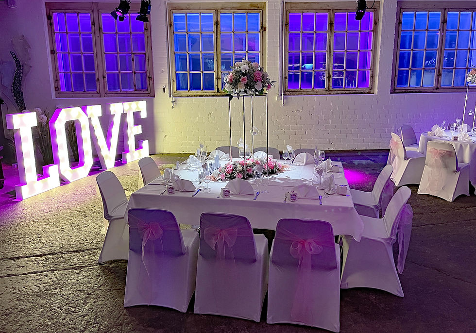 Hochzeitsdeko in lila beleuchtetem Raum. Tisch mit weißer Decke, rosa Blumen, Stühlen mit Schleifen. LED-Schriftzug LOVE leuchtet.