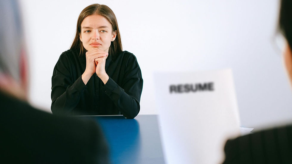 Woman in black shirt at a job interview, hands clasped, looking thoughtful. "Resume" is visible on a paper held by an interviewer.