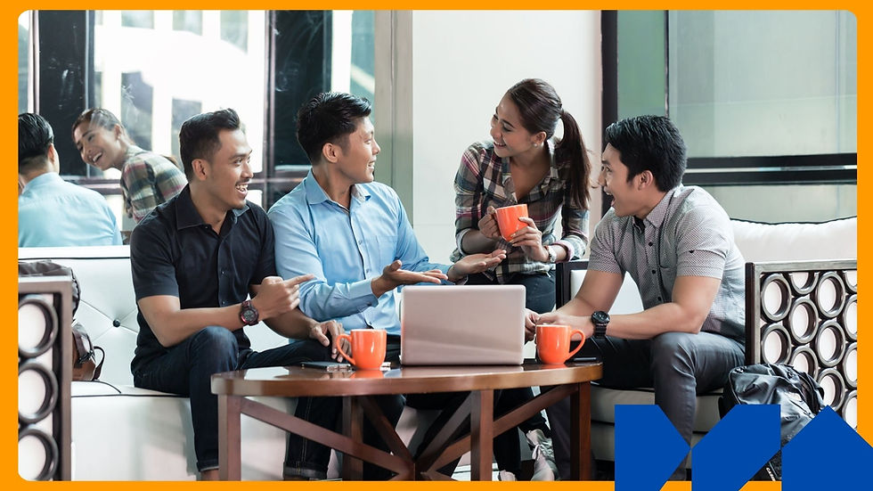 Four people smiling and chatting around a laptop in a bright room. Two hold orange mugs, creating a relaxed, friendly atmosphere.