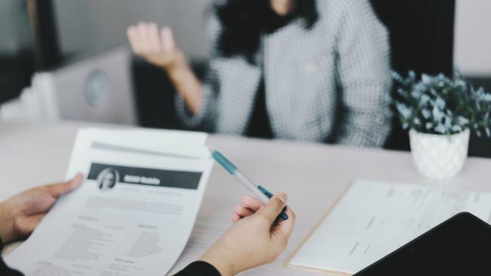 Person holding a resume and pen in an office setting; another person gestures. Blurred background with a plant, suggesting a job interview.
