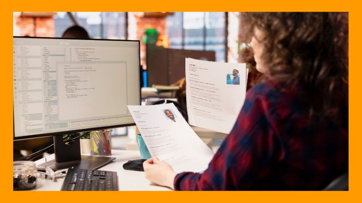 Person at a desk reviews resumes in an office with computer screens showing text. The atmosphere is focused and professional.