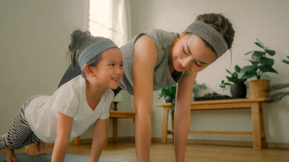 A woman and child smiling while doing push-ups on yoga mats in a bright room with plants and light wood floors, wearing matching headbands.