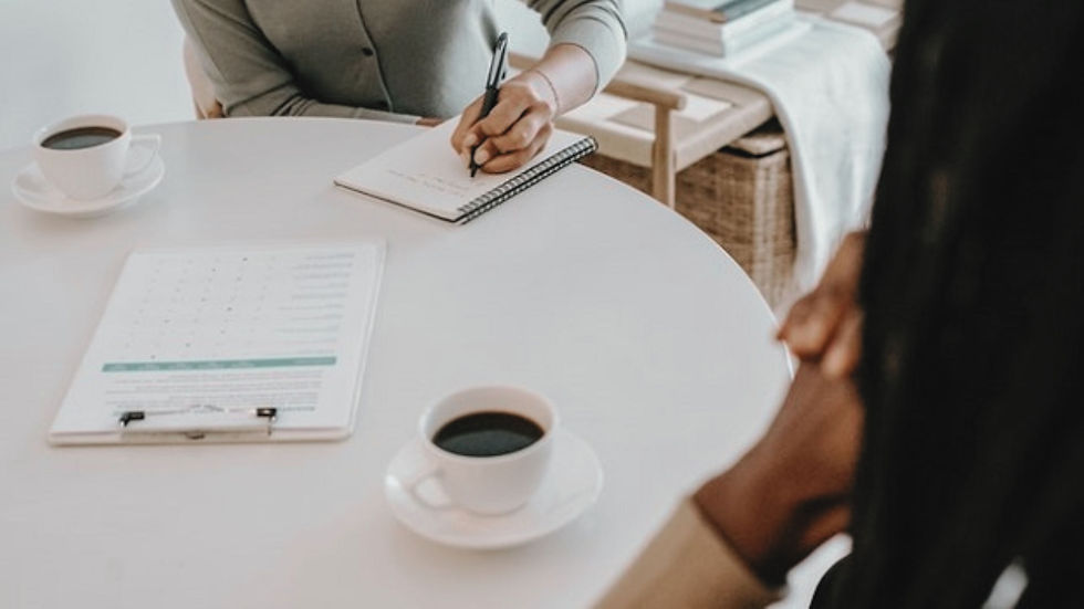 Two people at a white table with coffee cups. One writes in a notebook, while a clipboard with papers is visible. Neutral setting, calm mood.