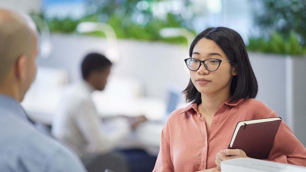 A woman in glasses holds a notebook, talking with a bald man in an office. Blurred background with plants and two people working at desks.