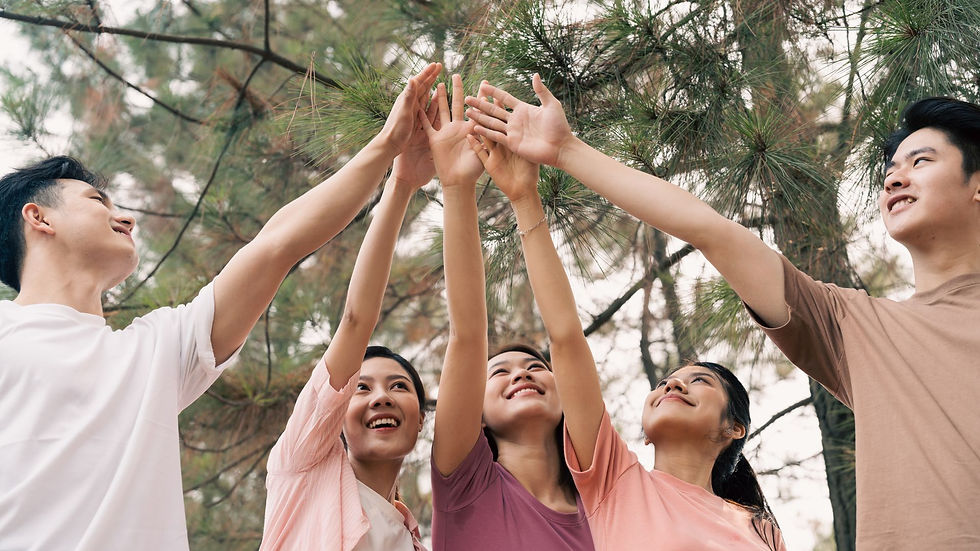 Five people smiling, reaching upward with hands together under trees. They wear pastel shirts, creating a joyful, outdoor scene.