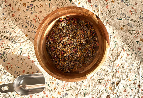 Kruiden die worden gebruikt voor de yoni-steam (o.a. calendula, bijvoet, rozenblaadjes, rozemarijn en kamille) in een houten kom, schepje er naast, op een katoenen tafelkleed met kruiden- en bloemetjesprint