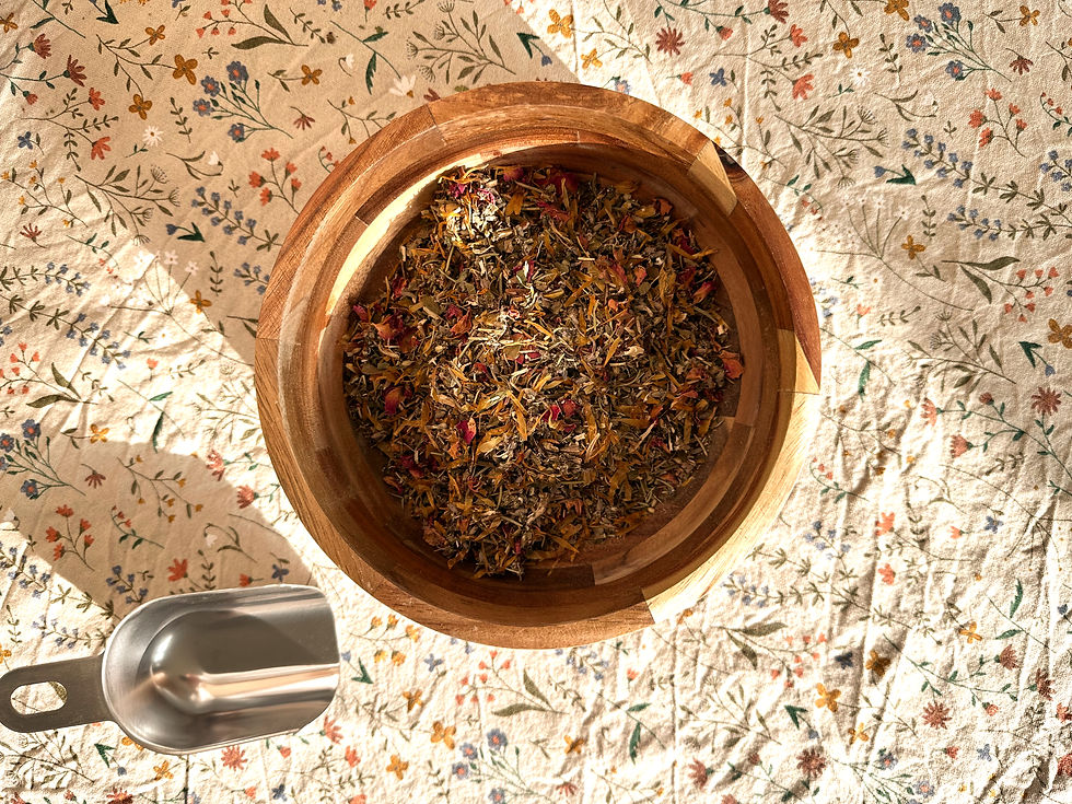 Kruiden die worden gebruikt voor de yoni-steam (o.a. calendula, bijvoet, rozenblaadjes, rozemarijn en kamille) in een houten kom, schepje er naast, op een katoenen tafelkleed met kruiden- en bloemetjesprint