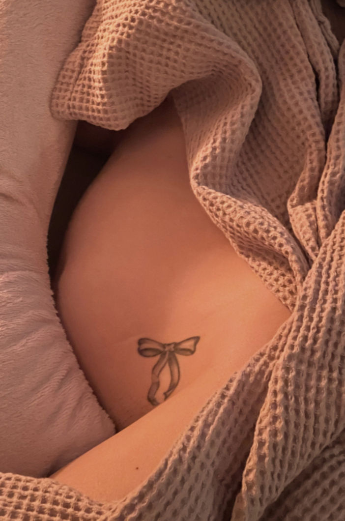 Zwangere buik die ingesnesteld ligt tussen een dekentje met wafelstructuur en een positioneringskussen, met zacht warm licht. Buik is ongeveer 18-20 weken zwangerschap en de vrouw heeft een tattoo van een strikje boven haar lies.