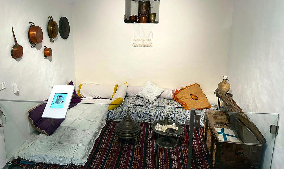 A small room with mattresses, pillows, and 19th-century Ottoman furnishings, including a purple rug, iron pots and pans, a wooden clothing chest, and a circular serving table
