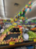A colorful grocery store produce section with apples, bananas, green apples, a weighing scale, and hanging piñatas in various shapes and colors above.