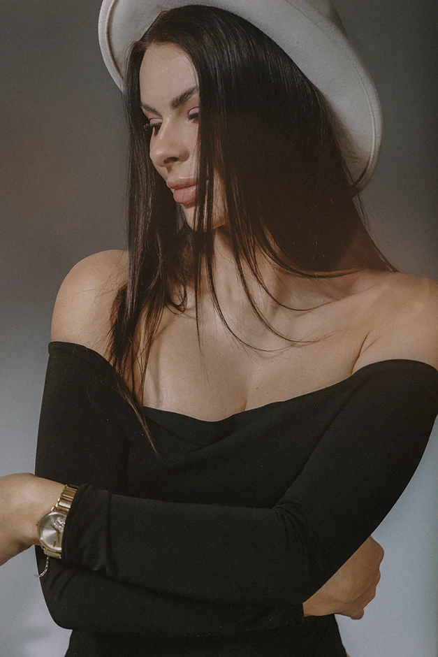 Zbliżenie na modelkę, która wyraża pewność siebie, ubrana w prosty, czarny top i spodnie, szare tło w kontrastowym świetle.