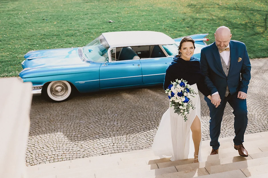 lenerowa ceremonia pod błękitnym niebem, wejście Pary Młodej wśród kwiatów, ikoniczny Cadillac Sedan DeVille przed pałacem, pałacowe wnętrza pełne blasku.