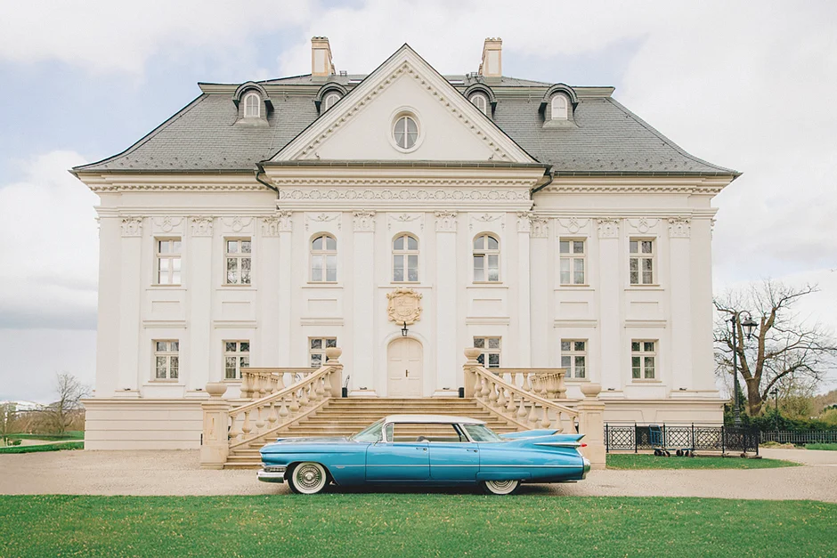 lenerowa ceremonia pod błękitnym niebem, wejście Pary Młodej wśród kwiatów, ikoniczny Cadillac Sedan DeVille przed pałacem, pałacowe wnętrza pełne blasku.