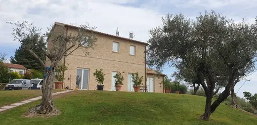 Casolare in pietra con pannelli solari sul tetto, circondato da ulivi e giardino curato, immerso nella campagna collinare sotto un cielo azzurro.
