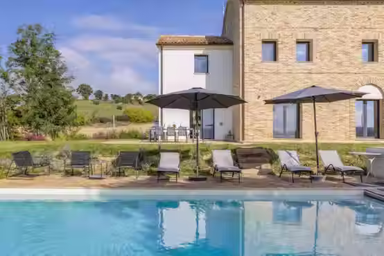 Vista esterna di una villa in pietra con piscina a sfioro, giardino curato e una pergola bianca con glicine che copre la zona pranzo all'aperto sotto un cielo azzurro con nuvole leggere.