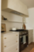 Modern Kitchen with Classic ElementsA modern kitchen with classic elements features sleek white cabinetry with brass knobs, a stainless steel oven, and minimalist open shelving. A plastered hood adds a touch of elegance, blending seamlessly with the overall design. A white pot sits on the stove, accompanied by decorative art and utensils, creating a warm and inviting cooking space.